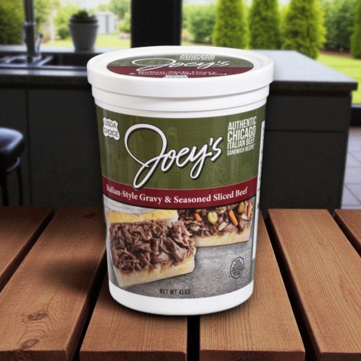 A container of Joey’s Italian Beef Sliced Beef & Vienna Beef Giardiniera Combo by Joey's Italian Beef rests on a wooden table, ideal for Chicago-style sandwiches, with a kitchen and greenery in the background.