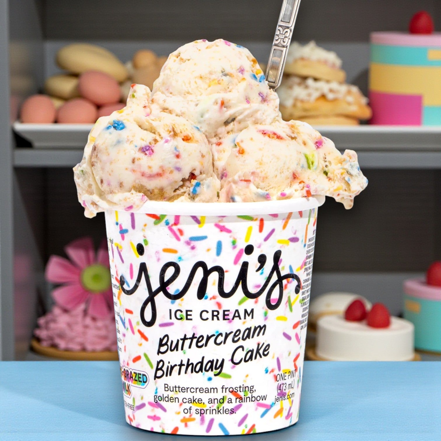 A container of Easy Lunches Premium Ice Cream Lovers Combo – 6 Total Pints, with three scoops, a spoon, and shelves filled with pastries and cakes in the background.
