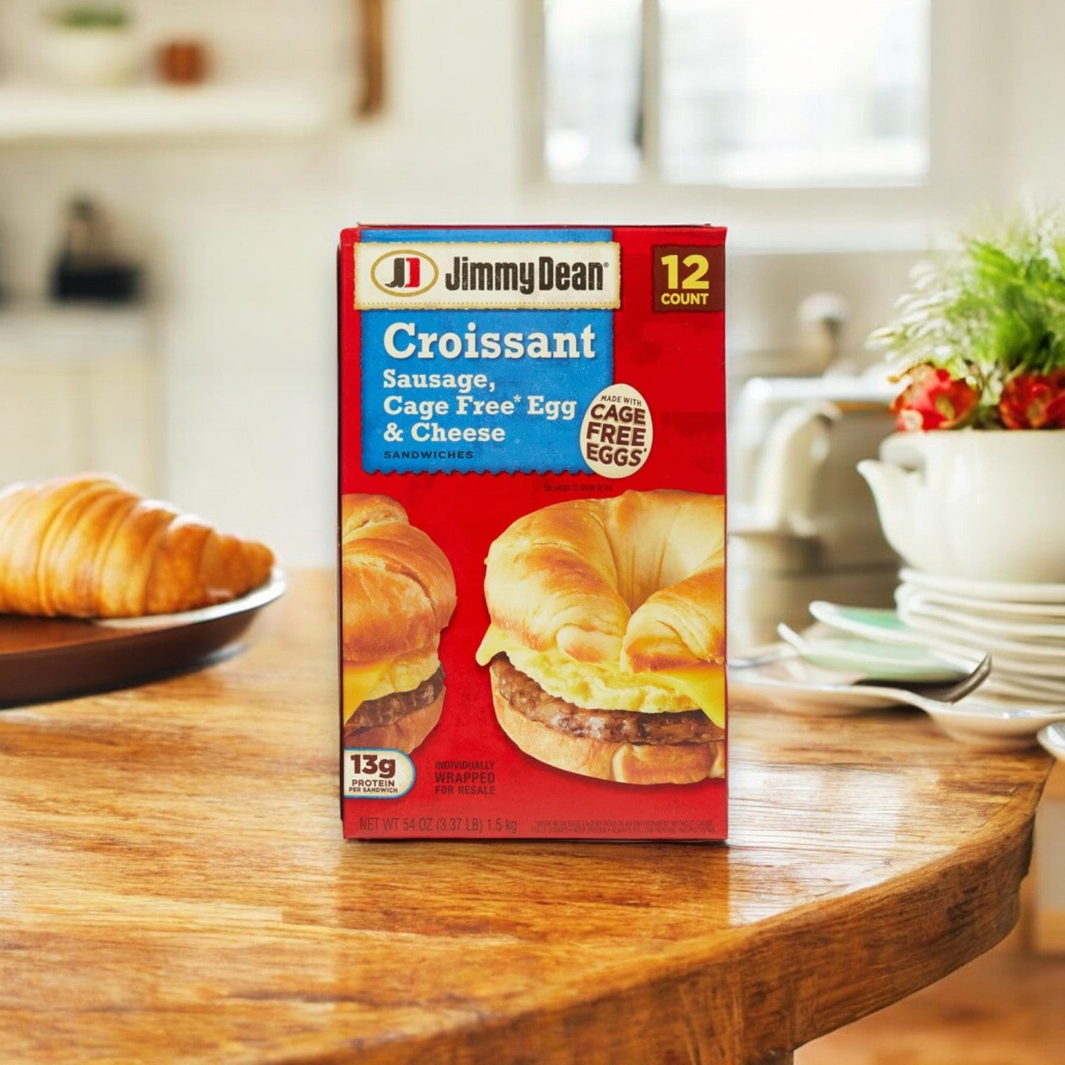 A box of Jimmy Dean Croissant Sandwich, Sausage, Cage Free Egg & Cheese, 12 ct - 1 Pack is displayed on a wooden table alongside a croissant and a plant in the background. Ideal for quick breakfast sandwiches that kickstart your day.