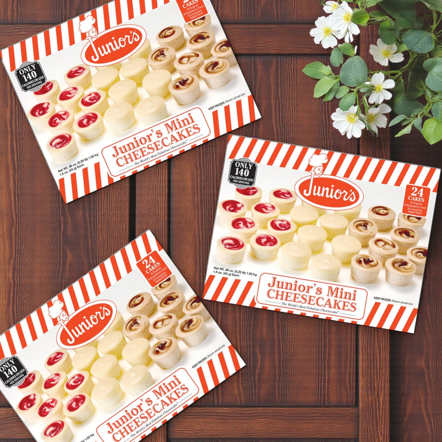 Three boxes of Juniors Mini Cheesecakes – Variety Pack, 1.5 oz, 24 Count (3 Packs) are displayed on a wooden surface beside a small potted white-flower plant.