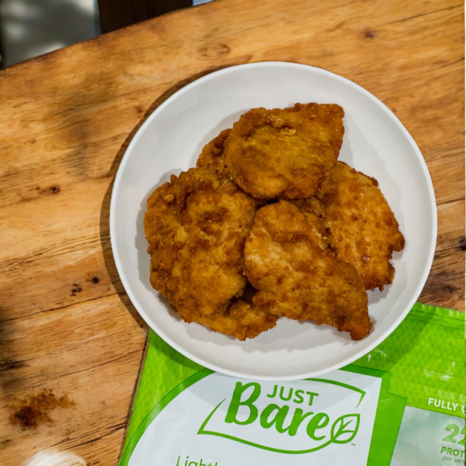 Five pieces of protein-packed, Just Bare Lightly Breaded Chicken Breast are served on a white plate, placed on a wooden surface with green Just Bare packaging partially visible beside it.