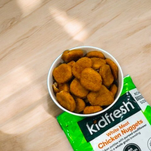 A bowl filled with Kidfresh Frozen Chicken Nuggets - 16.35 oz, made from white-meat chicken, sits on a wooden surface next to a green Kidfresh packaging bag. Perfect for picky eaters, these nuggets contain hidden veggies for added nutrition.