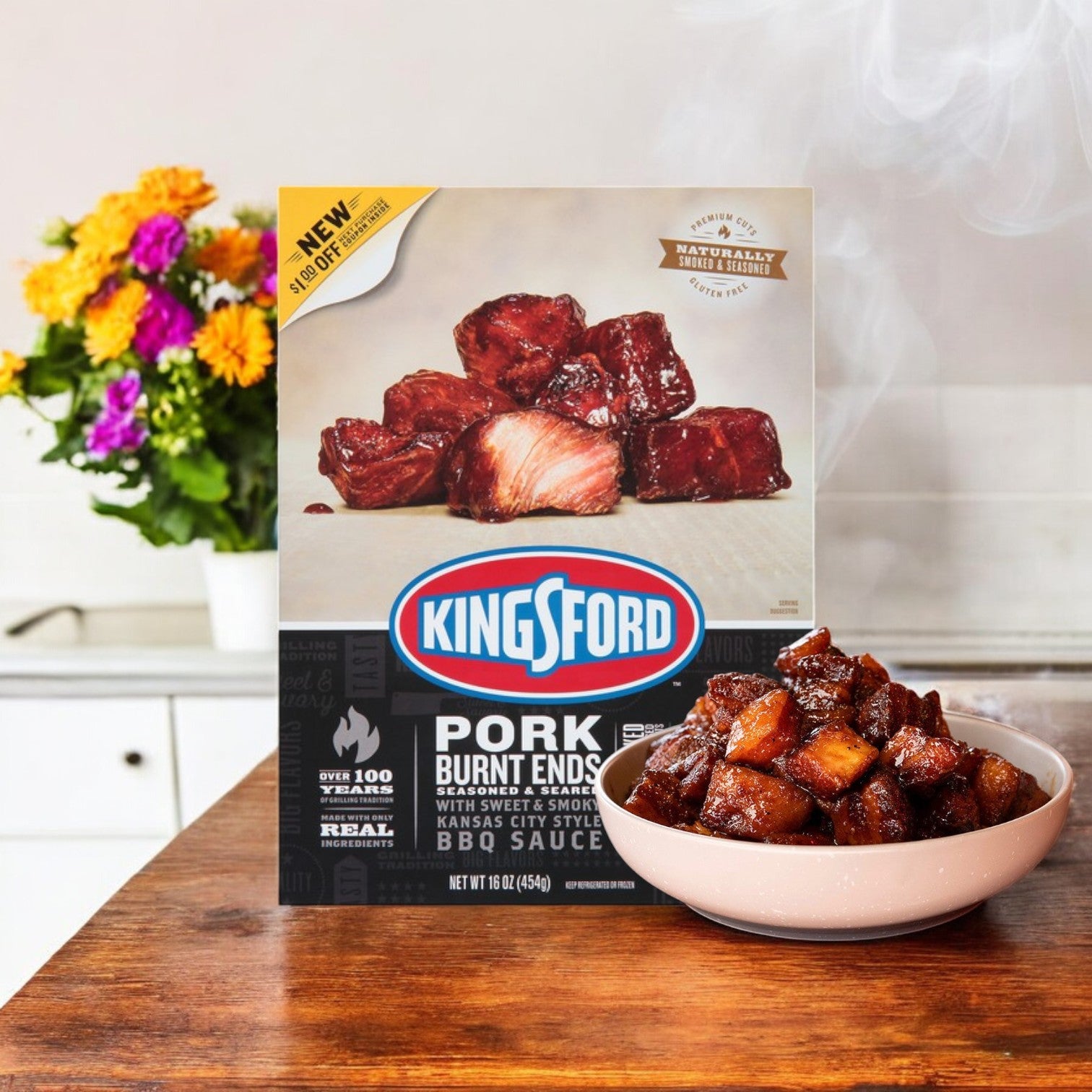 A box of Kingsford Pork Burnt Ends – Seasoned & Seared, 16 oz, rests on the kitchen counter beside a bowl of smoky BBQ goodness. Flowers frame this savory display, adding elegance.