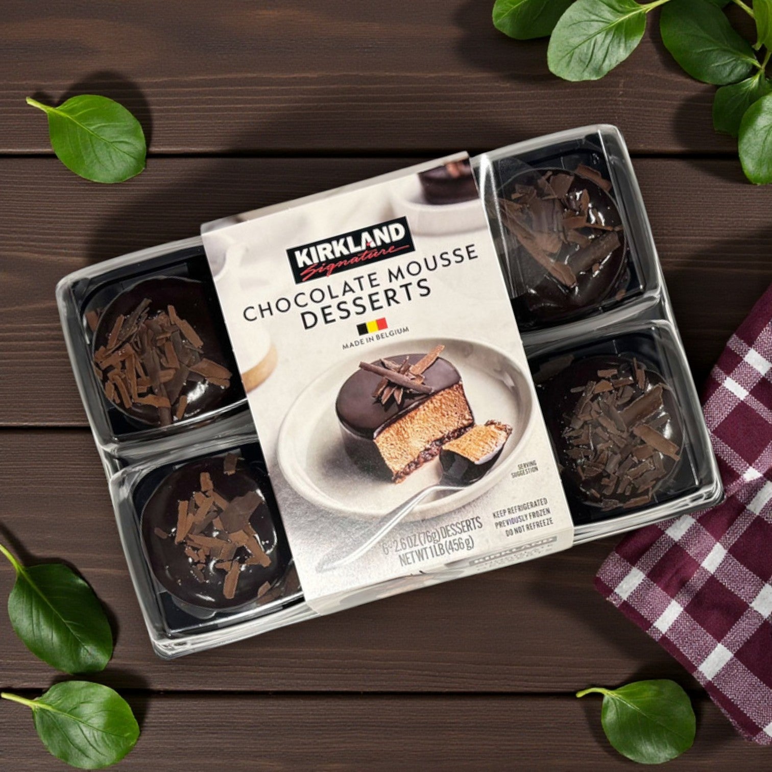 Kirkland Signature Chocolate Mousse Desserts, 2.6 oz, 6-Count feature six individual premium cocoa treats in a clear container, set on a dark wooden surface with green leaves and a checked napkin nearby.