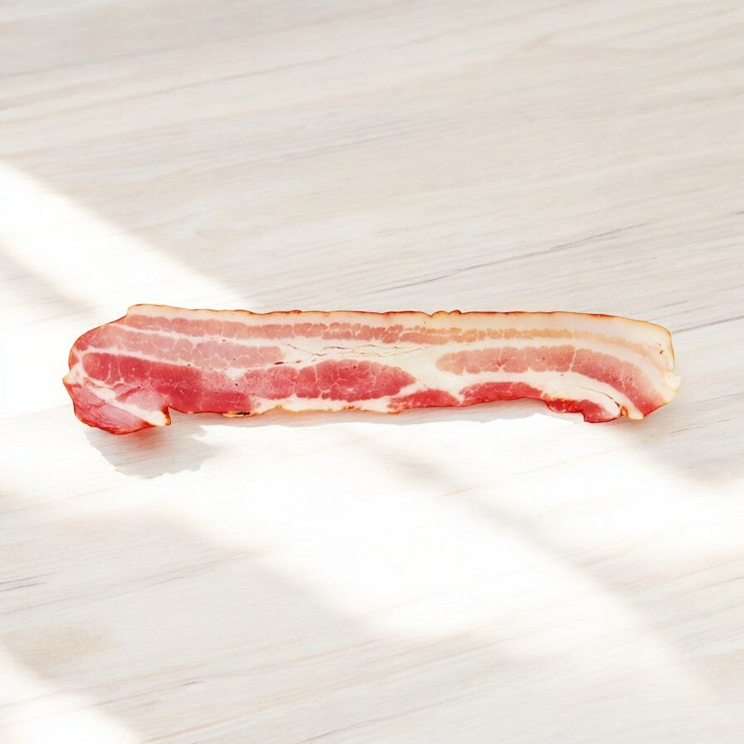 A raw strip of Easy Lunches Kirkland Signature Thick Sliced Bacon (1.5 lbs, 2 count packs, 3 packs—9 lbs total) rests on a light wooden surface with diagonal sunlight casting shadows.