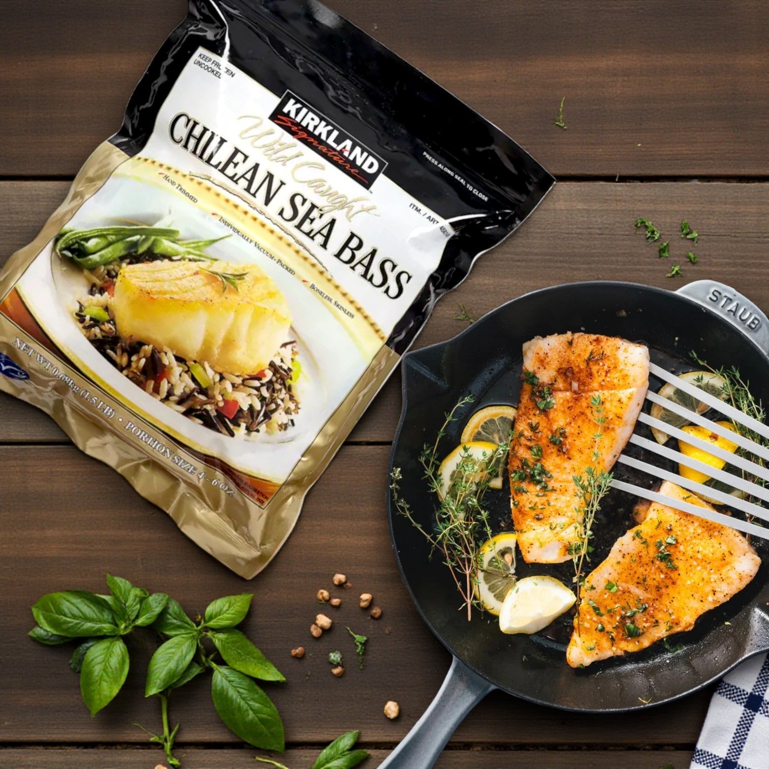 A package of Kirkland Signature Wild Chilean Sea Bass – 4–6 oz Portions (1.5 lbs Total) sits next to a skillet with two cooked fillets, lemon slices, and herbs on a wooden surface with basil and peppercorns.