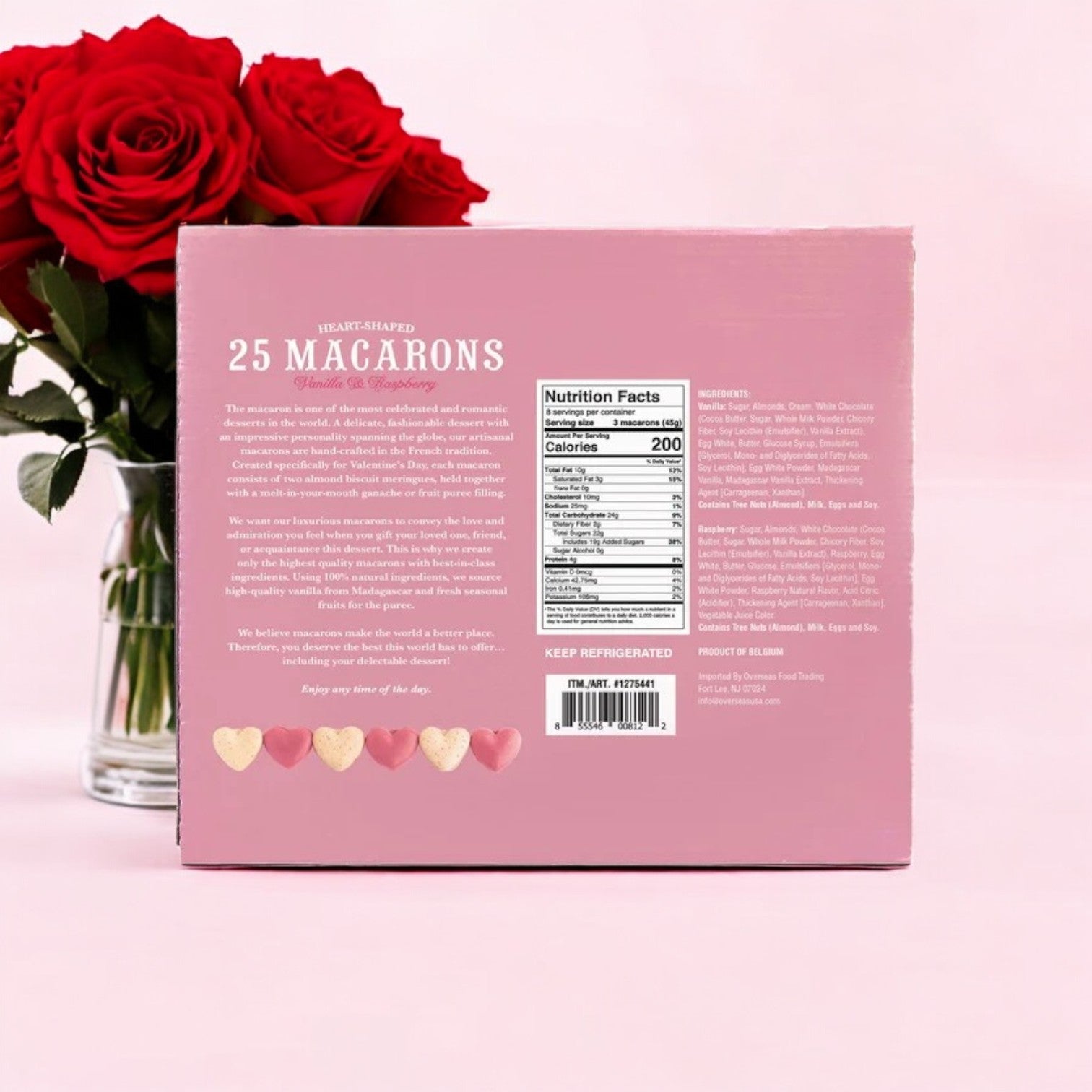 A pink box of La Chic Patissier Heart Macaron – 25-Count, 13.2 oz., showcasing premium quality and diverse flavors, is elegantly nestled beside a vase of red roses. Each macaron includes a nutrition facts label to ensure you savor every bite confidently.