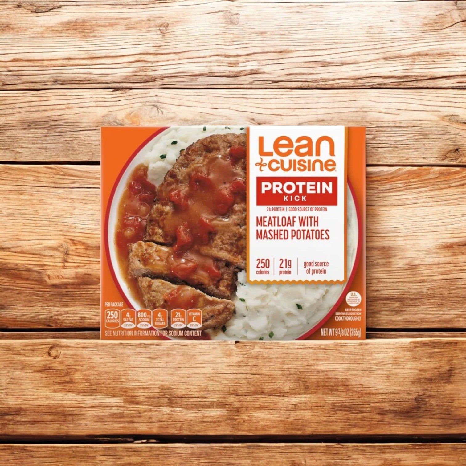 A box of Lean Cuisine Meatloaf With Mashed Potatoes Frozen Entrée, weighing 9.375 oz., features 250 calories, 21g of protein, and no preservatives. Nestled beside it is a delectable Mac and Cheese made with Vermont White Cheddar. Both packages are displayed on a wooden surface.