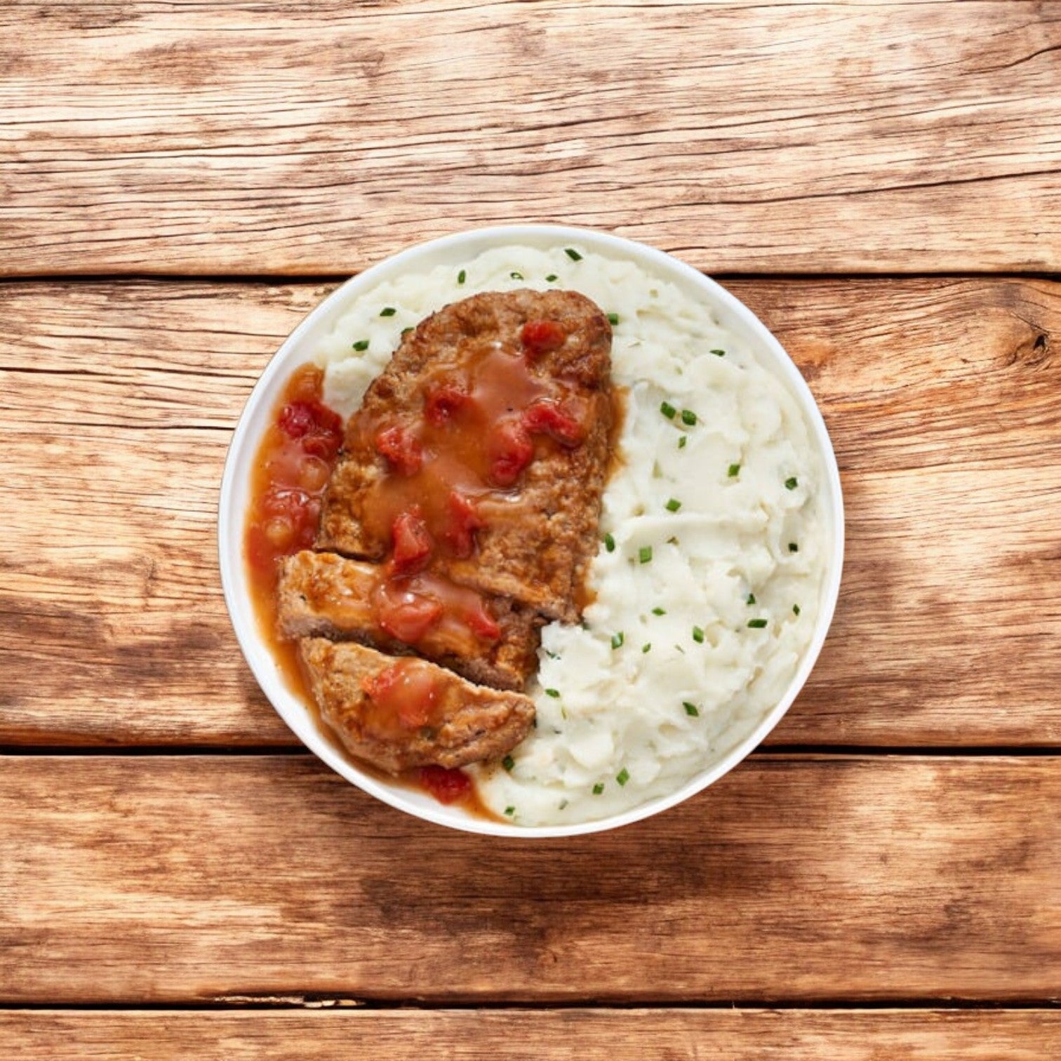 A white bowl containing Lean Cuisine Meatloaf With Mashed Potatoes Frozen Entrée, garnished with chives and topped with gravy, sits on a wooden surface next to a steaming plate of Vermont White Cheddar Mac and Cheese.