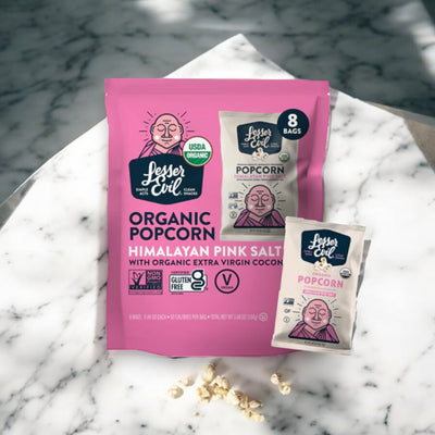 A package of LesserEvil Himalayan Pink Salt popcorn is displayed on a marble surface alongside one smaller individual serving bag. The packaging highlights various certifications, showcasing it as a vegan-friendly, healthy snack option.