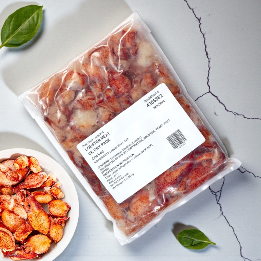 A sealed Easy Lunches bag of Maine Lobster Meat – Claw & Knuckle, 2 lbs., with a printed label rests on a cracked gray surface beside a plate of lobster meat and green leaves.