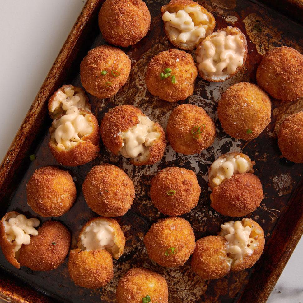 A baking sheet filled with golden-brown, round croquettes, some of which are split open to reveal a creamy Feel Good Foods Cheese Mac & Cheese filling from a 1 Box - 8 oz package.