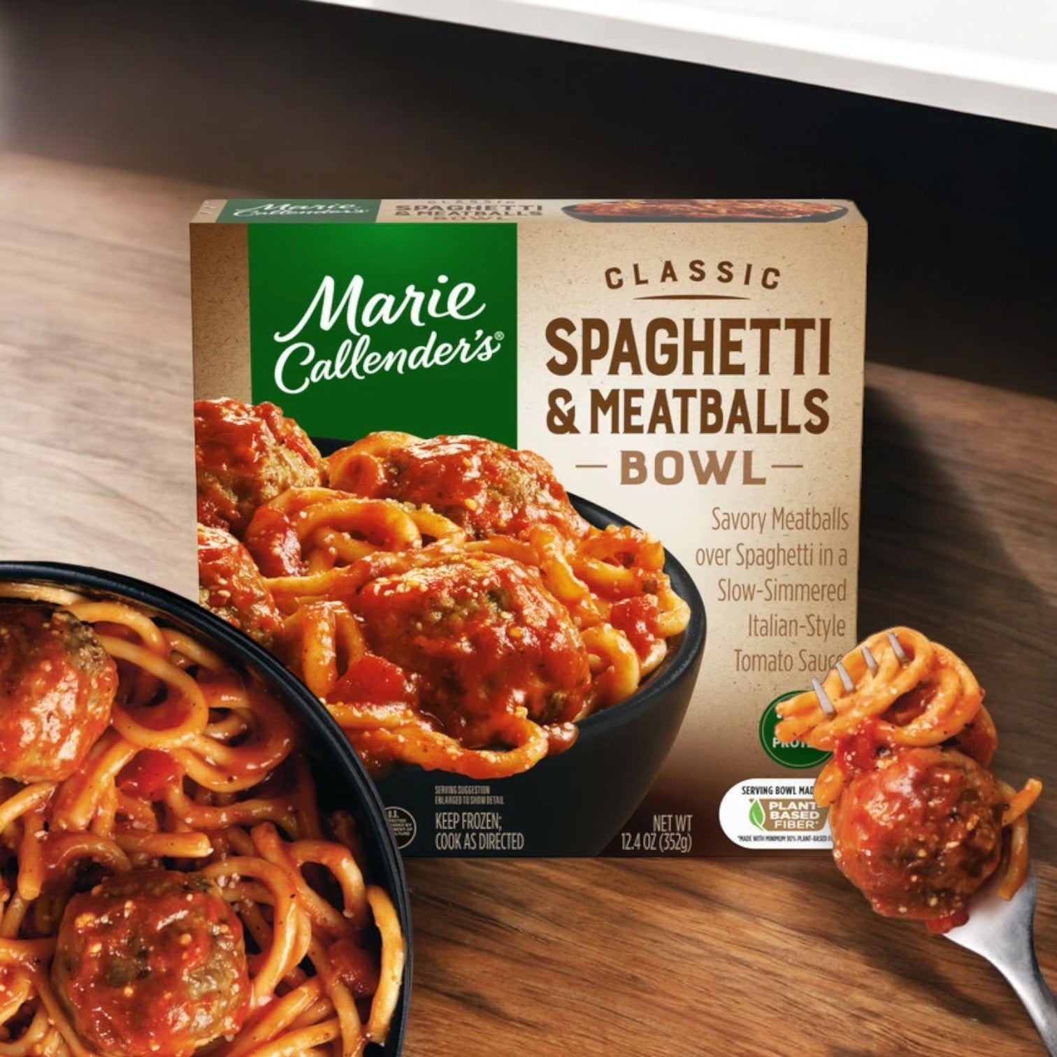 A box of Marie Callender's Classic Spaghetti and Meatballs Bowl, 12.4 oz. - 1 Count, a comforting favorite from the brand Marie Callender's, sits beside a bowl of spaghetti and meatballs on a wooden surface. The packaging showcases an enticing image of this frozen Italian dish along with product details.