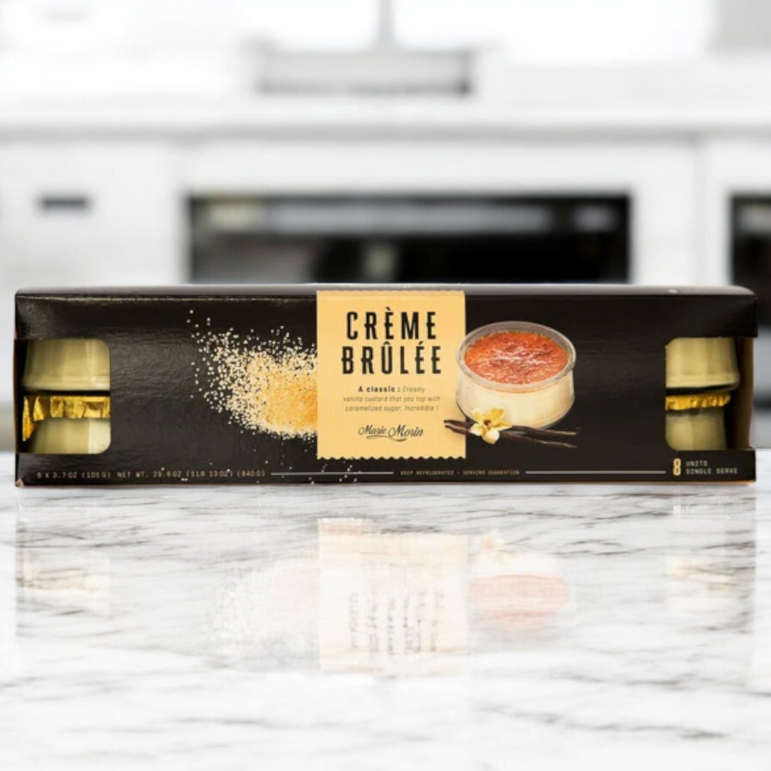 A box of Creme Brulee - Marie Morin on a marble countertop, featuring eight 3.7oz ready-to-serve containers adorned with an enticing dessert image and vanilla accents on the packaging—offering a premium dessert experience that embodies the essence of this classic French treat by CRÈME BRULEE - MARIE MORIN.