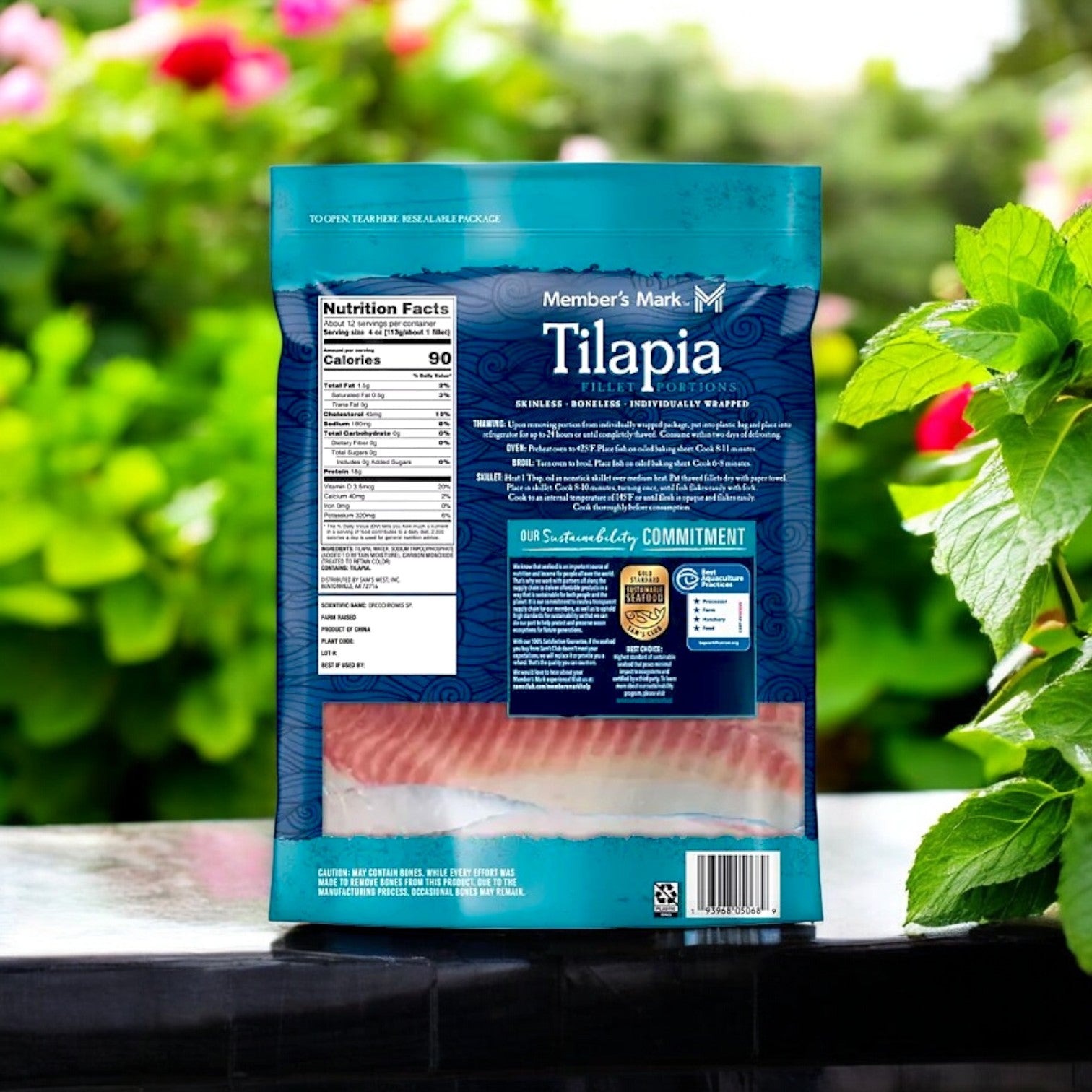 A package of Member's Mark Farm-Raised Tilapia Fillet Portions (9 lbs., skinless, boneless, frozen) sits on a table showcasing its nutrition and sustainability info, with greenery and pink flowers in the background, highlighting this lean protein option.