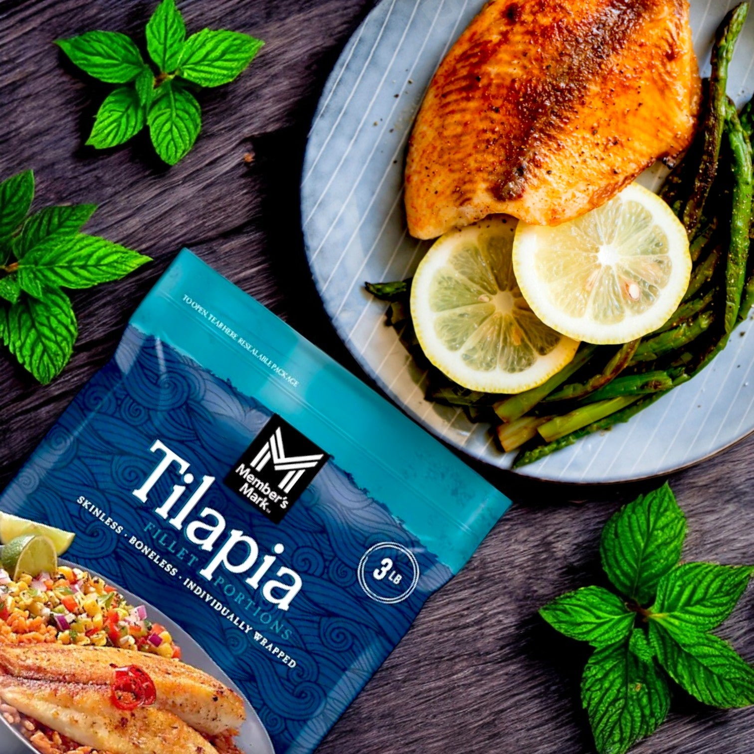 A Member's Mark Farm-Raised Tilapia Fillet Portion is displayed next to a prepared dish of cooked tilapia with lemon slices and green beans. Green leaves scatter across a wooden surface, highlighting this delicious source of lean protein.