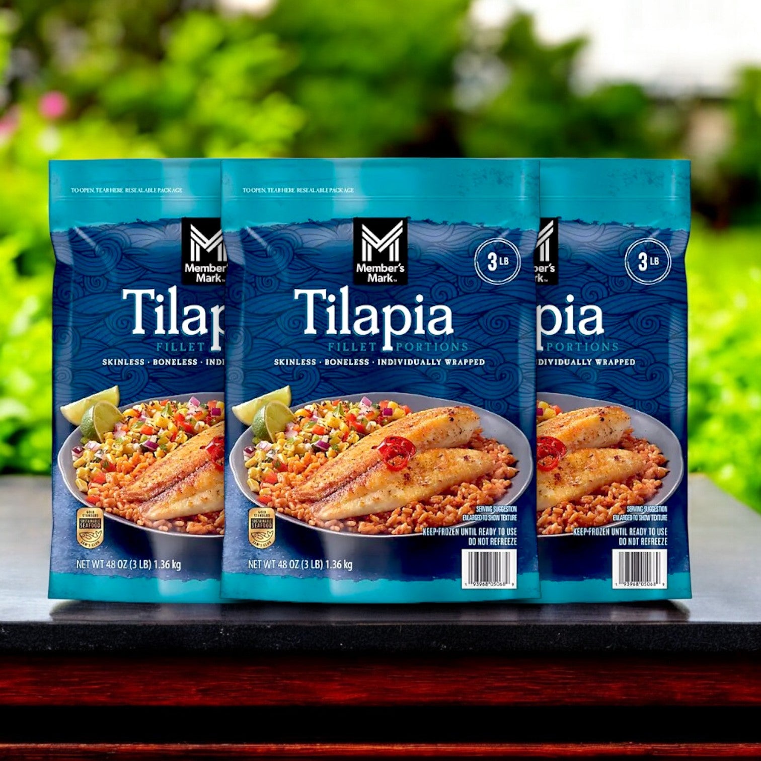 Three packs of Member's Mark Farm-Raised Tilapia Fillet Portions are displayed outdoors with lime and rice garnish, showcasing this skinless, boneless, frozen product as a delicious lean protein source.