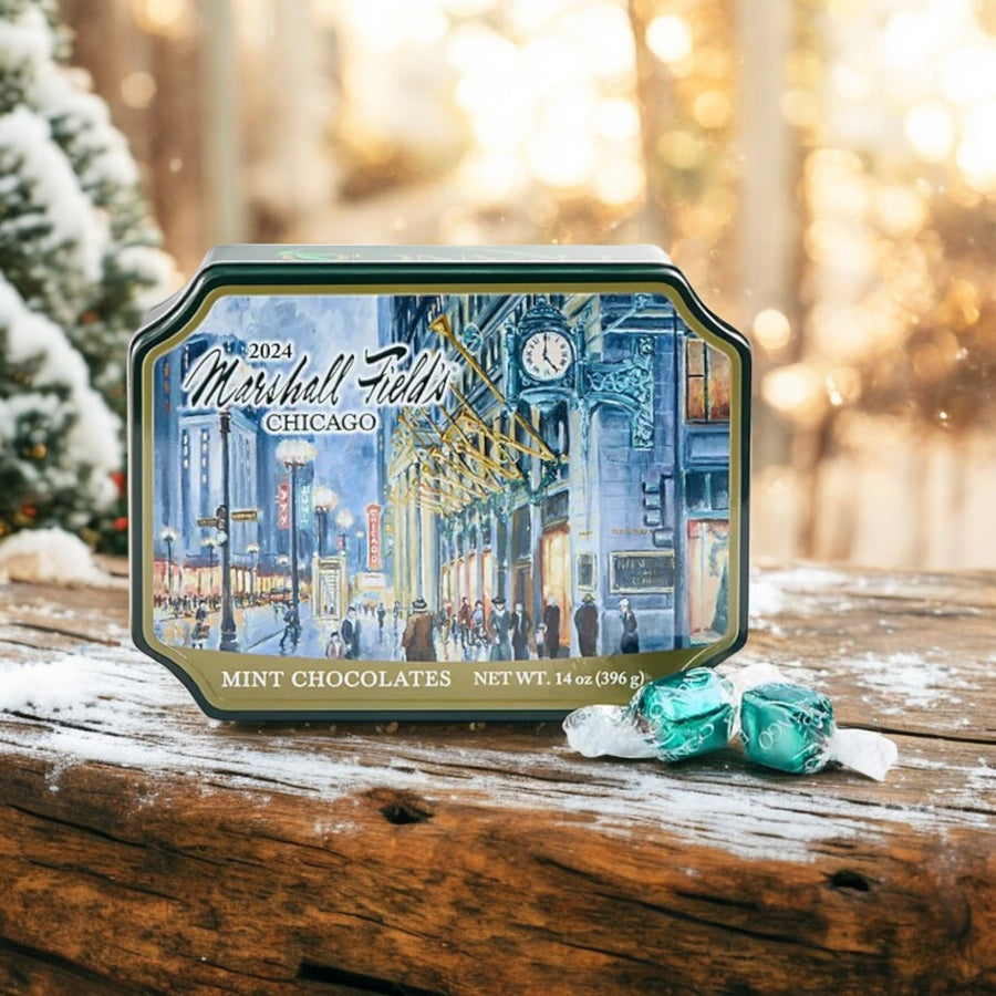 A Garrett Marshall Field's Holiday Clock Tin – Milk Mint Chocolates (14 oz) rests on a wooden surface with two unwrapped candies nearby, set against a snowy, festive backdrop—ideal for holiday gifting.