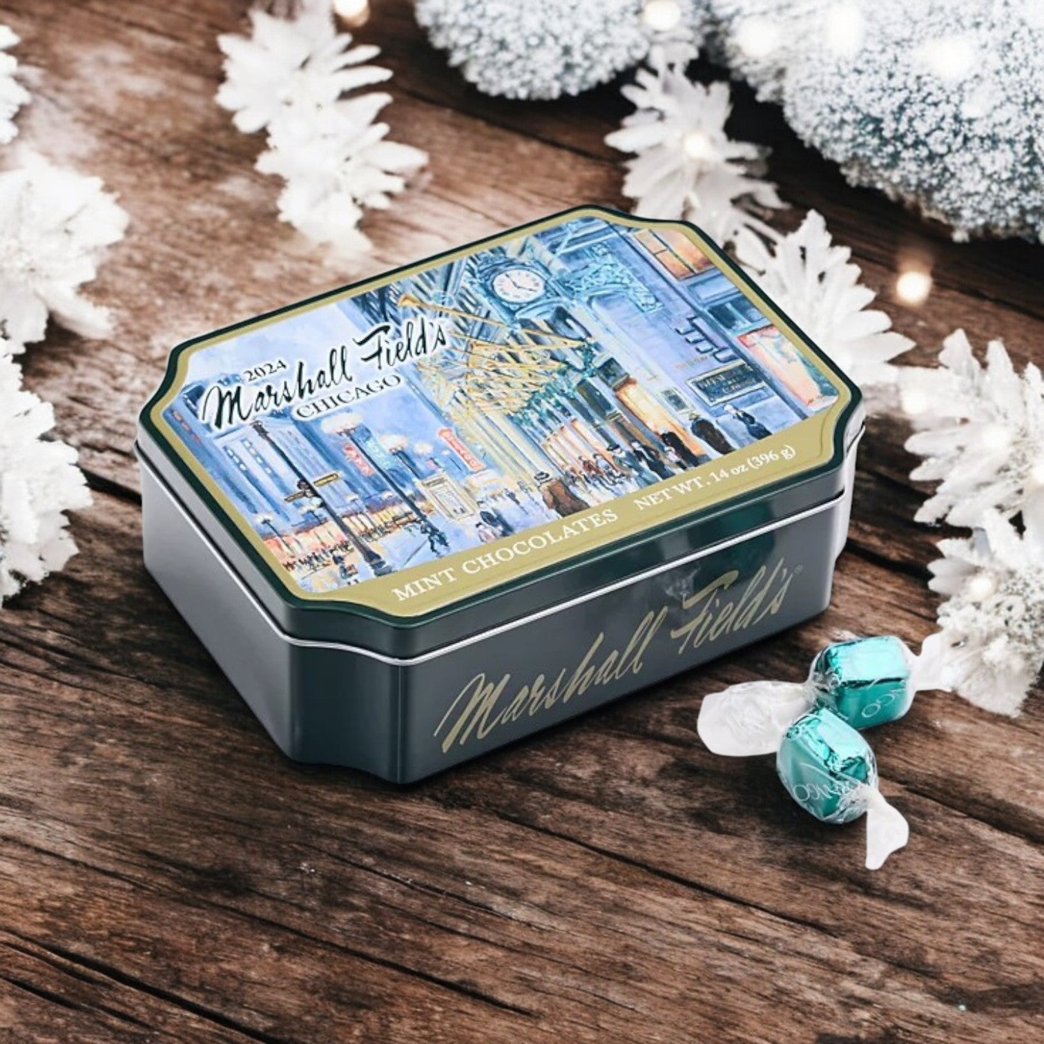 A Garrett Marshall Field's Holiday Clock Tin, containing 14 oz of Milk Mint Chocolates, is displayed on a wooden surface adorned with snowflakes and illuminated string lights. This collectible keepsake brings festivity to any setting.