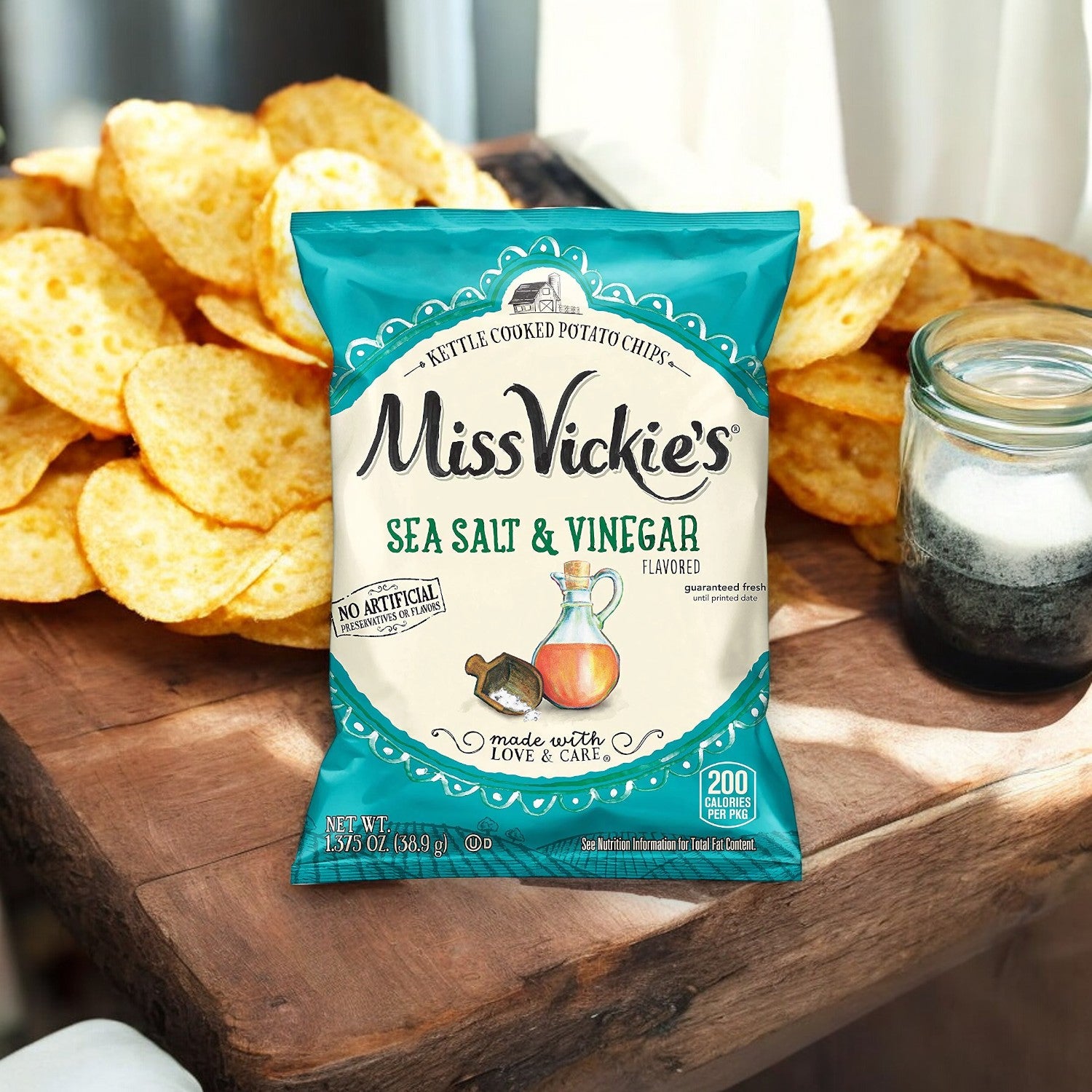 A 1.375 oz bag of Miss Vickie's Sea Salt & Vinegar Flavored Kettle Cooked Potato Chips is placed on a wooden table, surrounded by loose potato chips, with a jar of salt in the background, promising a natural snacking experience with its tangy vinegar seasoning.