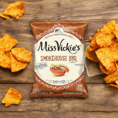 A 1.375 oz bag of Miss Vickie's Smokehouse BBQ flavored kettle cooked potato chips, proudly featuring no artificial flavors, with several crisps scattered on the wooden surface around the bag.