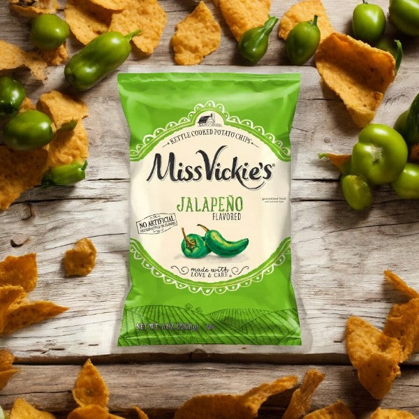 A bag of Miss Vickie's Jalapeno 1-1.375 oz kettle cooked potato chips, renowned for their spicy kick and absence of artificial flavors, is surrounded by whole jalapenos and broken pieces of chips on a wooden surface.