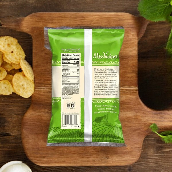 Back view of a green bag of Miss Vickie's Jalapeno 1-1.375 oz potato chips on a wooden surface, with visible nutrition facts label. Chips are scattered on the left side and a bunch of green leaves is in the top right corner. The bag proudly states "no artificial flavors.