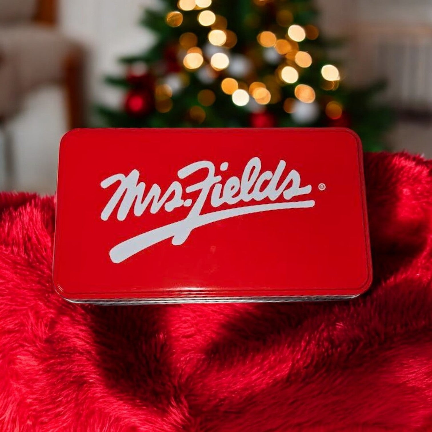 A red Mrs. Fields Signature Tin filled with their Nibblers Bite-Sized Assorted Cookies sits on a crimson blanket, as a softly twinkling Christmas tree fades into the background.