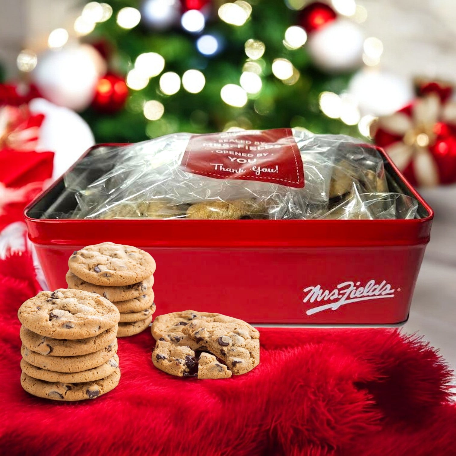 A "Mrs. Fields" signature tin, containing mouthwatering Nibblers bite-sized milk chocolate chip cookies, is displayed on a red cloth. A stack of these delectable 24 treats sits in the foreground, with a beautifully decorated Christmas tree casting a festive blur in the background.