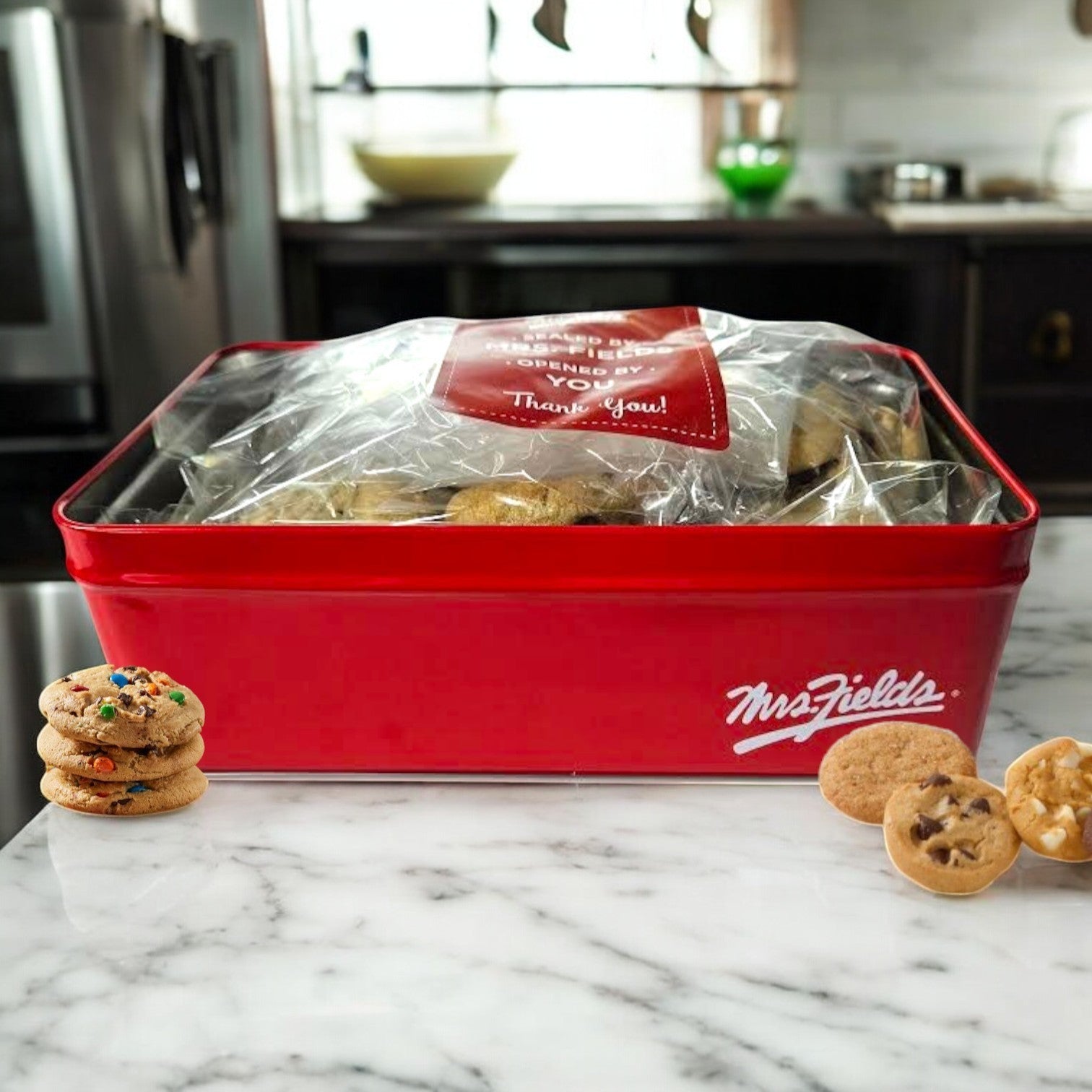 The red Mrs. Fields Signature Tin sits on a marble countertop, filled with bite-sized assorted Nibblers cookies. Three cookies are displayed in front: one with candy and two classic chocolate chip varieties.