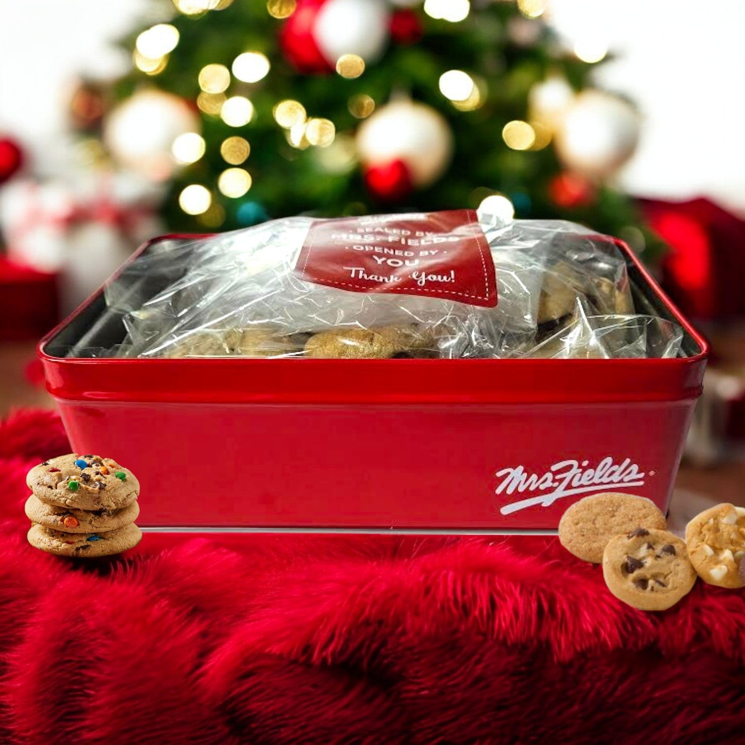 A Signature Tin of Mrs. Fields Nibblers Bite-Sized Assorted Cookies, containing 24 delightful treats, is encased in clear packaging. It sits atop a red furry cloth, with softly blurred holiday decorations providing a festive background.