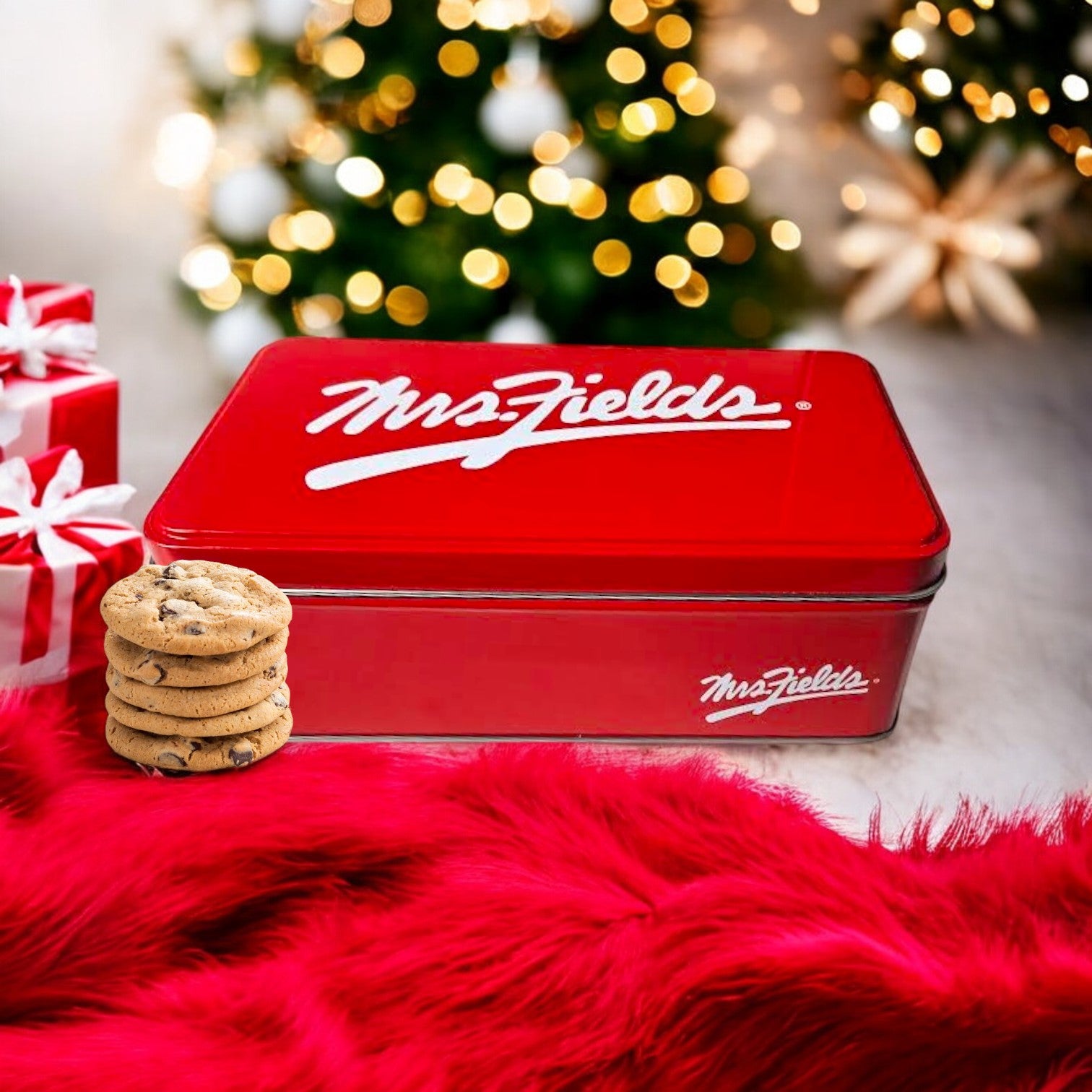 A Mrs. Fields Signature Tin, featuring Nibblers Bite-Sized Milk Chocolate Chip Cookies with 24 delicious treats, rests on a red cloth. Nearby, cookies are neatly stacked while Christmas lights twinkle around them and wrapped gifts add to the festive atmosphere.