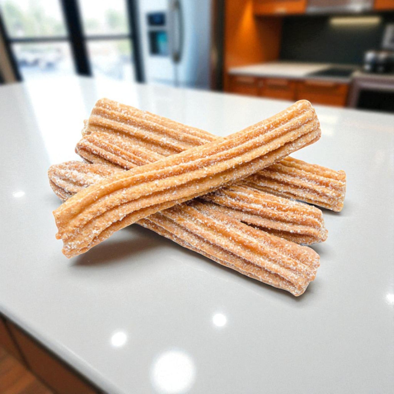 Three Sabor Nuestro Frozen Cajeta-Filled Churros (10", 50 ct) are stacked on a white countertop in a modern kitchen with a blurred background, showcasing this delicious take on classic Mexican desserts.