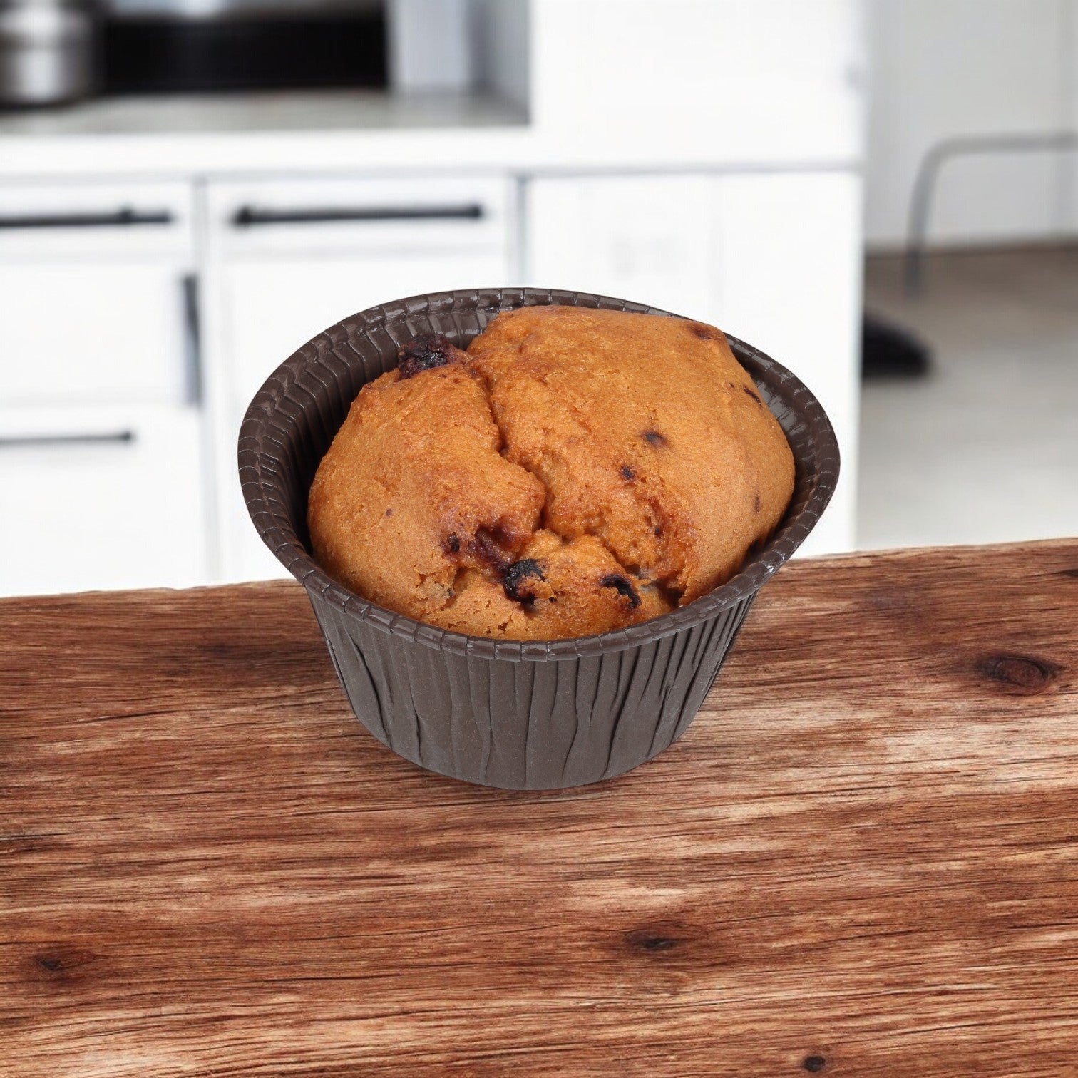 An individually-wrapped blueberry muffin in a brown paper cup is placed on a wooden table with a kitchen in the background, reminiscent of Udi's inviting bakery where each treat is crafted for freshness.