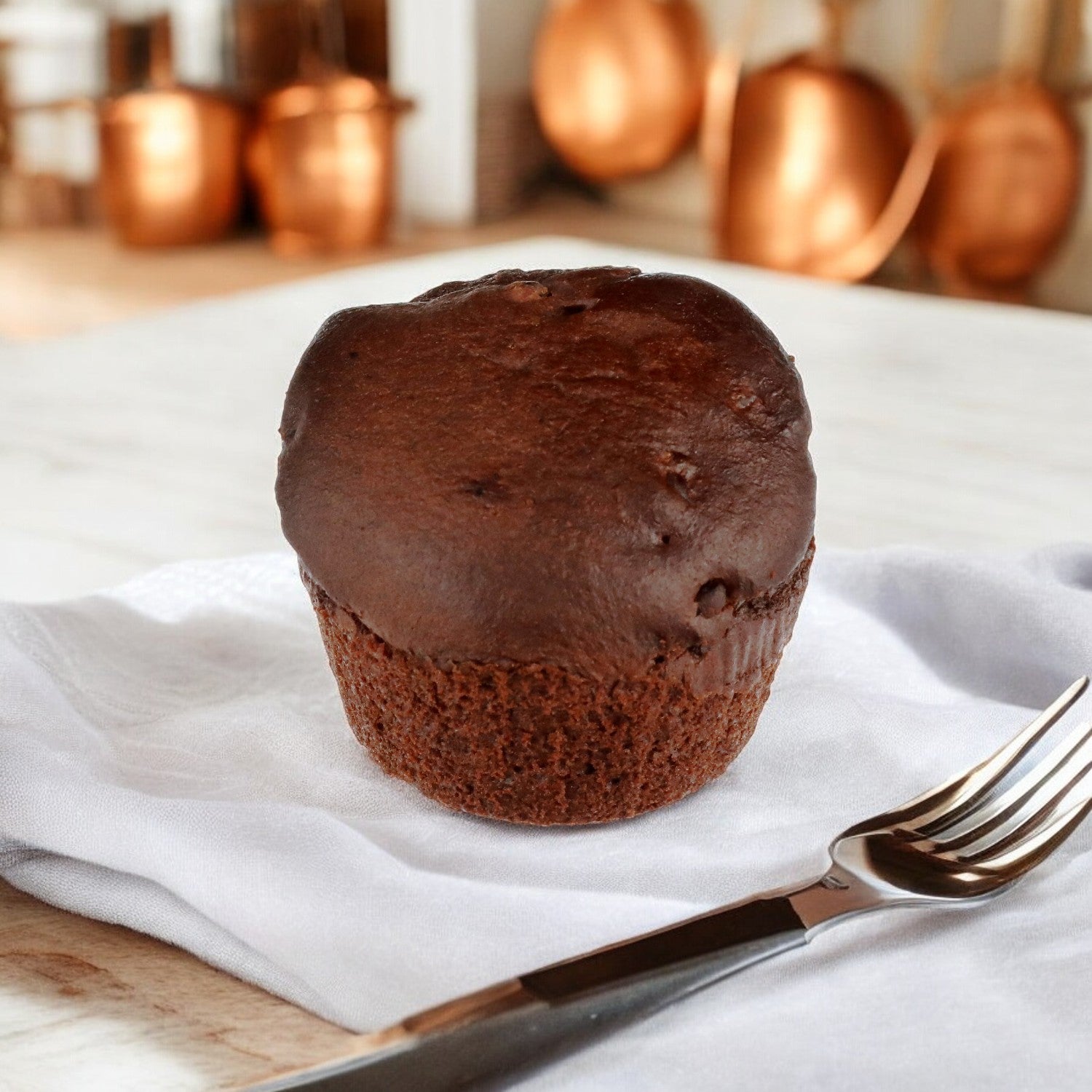 An individually wrapped Udi's Double Chocolate Gluten-Free Muffin, weighing 3 oz., sits atop a white napkin with a fork nearby, as blurred copper pots in the background accentuate its rich allure.