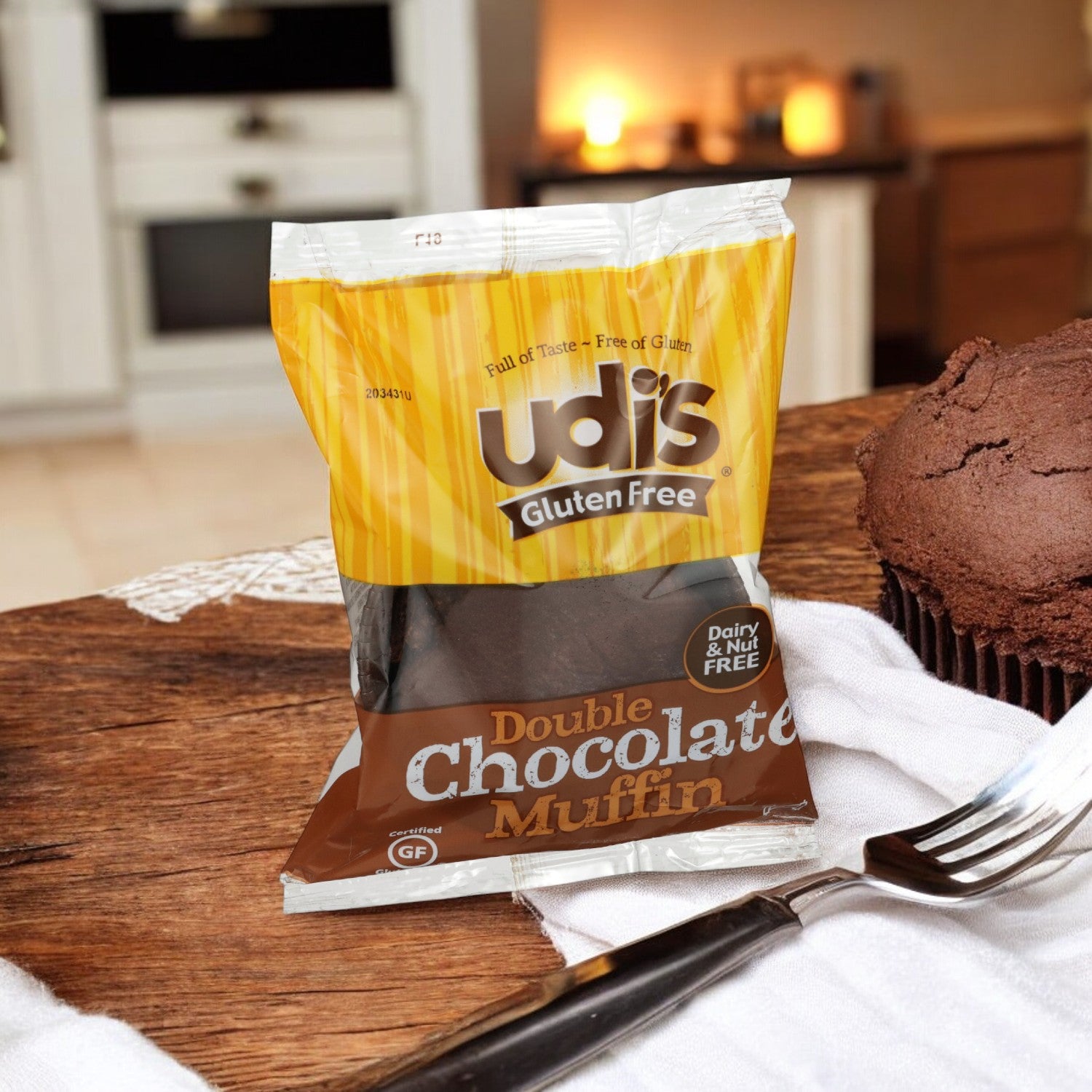 A package of Udi's Muffin Chocolate Double Gluten Free sits on a wooden table, accompanied by an unwrapped muffin and a fork. The cozy kitchen setting in the background emphasizes the convenience of these individually wrapped 3 oz. treats, perfect for immediate enjoyment.