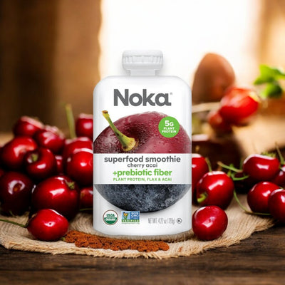 A pouch of Noka Organic Cherry Acai Smoothie - 4.22 oz - 1 Count sits on a wooden surface, surrounded by fresh cherries and smoothie powder. This organic, vegan blend from Noka is packed with antioxidants and prebiotic fiber, offering a delicious boost to your day.