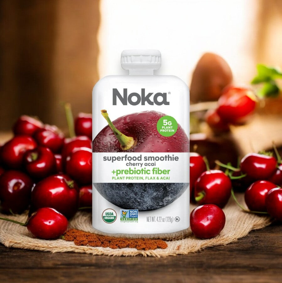 A pouch of Noka Organic Cherry Acai Smoothie - 4.22 oz - 1 Count sits on a wooden surface, surrounded by fresh cherries and smoothie powder. This organic, vegan blend from Noka is packed with antioxidants and prebiotic fiber, offering a delicious boost to your day.