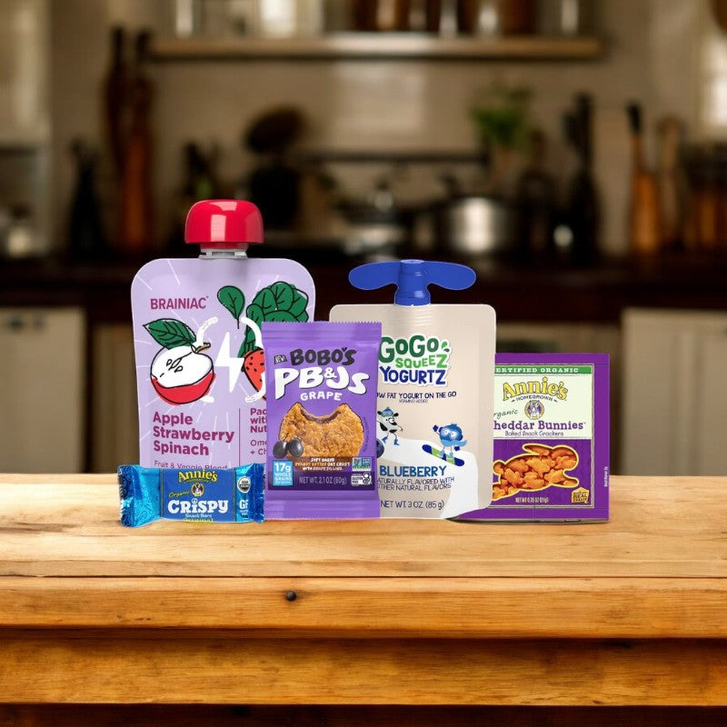 An assortment of wholesome snack products, like Bobo's PB&J's Grape gluten-free oat bar, organic apple strawberry spinach puree, blueberry yogurt, crispy rice treat, and cheddar bunnies are elegantly displayed on a wooden surface.