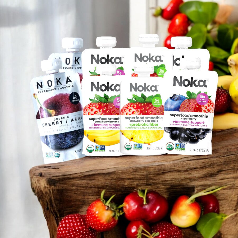 The Noka Variety Pack, featuring 4 flavors with 2 packs of each for a total of 8 packs, is displayed on a wooden surface surrounded by an assortment of fruits including bananas, strawberries, and cherries.