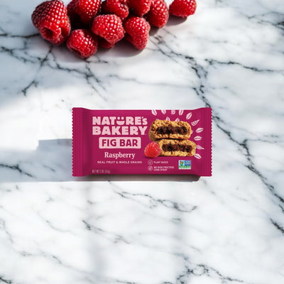 A single 2 oz Nature's Bakery Raspberry Fig Bar from Nature's Bakery sits on a marble surface, crafted with whole wheat flour and fig paste, with fresh raspberries in the background.