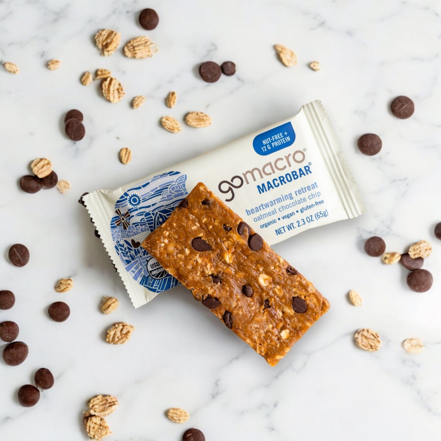 A GoMacro Oatmeal Chocolate Chip MacroBar-2.3 oz, with oats and chocolate pieces scattered around it, placed on a light marble background. This gluten-free, plant-based treat from GoMacro is perfect for a nourishing snack anytime.