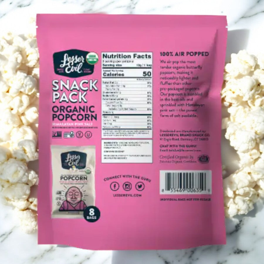 A bag of LesserEvil Himalayan Pink Salt popcorn snack packs sits on a marble surface, surrounded by popped popcorn. The back of the bag is visible, showcasing nutritional facts and product information, emphasizing its vegan-friendly ingredients. Ideal for anyone in search of a healthy snack option.