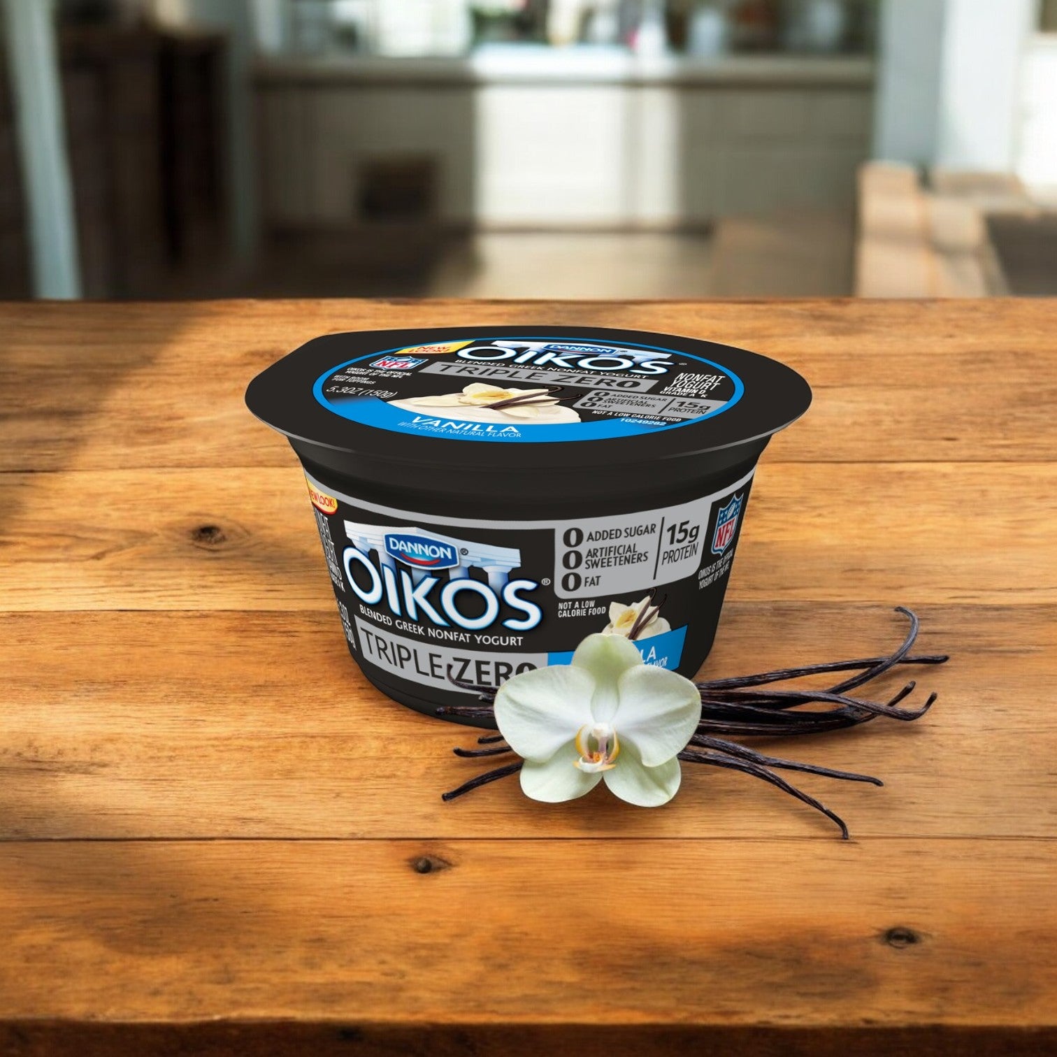 A container of Dannon's Oikos Triple Zero Vanilla Yogurt, 5.3 oz. from a case of 12, known for being high in protein and containing no added sugar, sits on a wooden table next to a white orchid flower and vanilla beans. The background features a blurred kitchen setting.