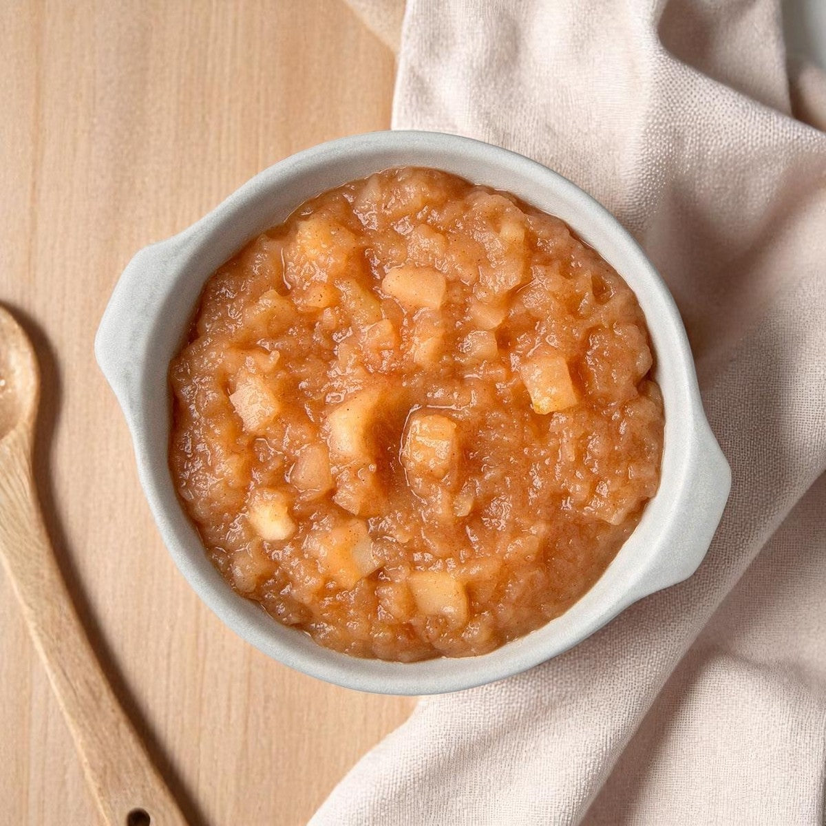 A white bowl of Sweet Street Scoopable Apple Compote sits on a light wooden surface beside a beige cloth and wooden spoon—perfect as a delicious dessert topping. Available in 3 bulk tubs (102 oz each) by Sweet Street.