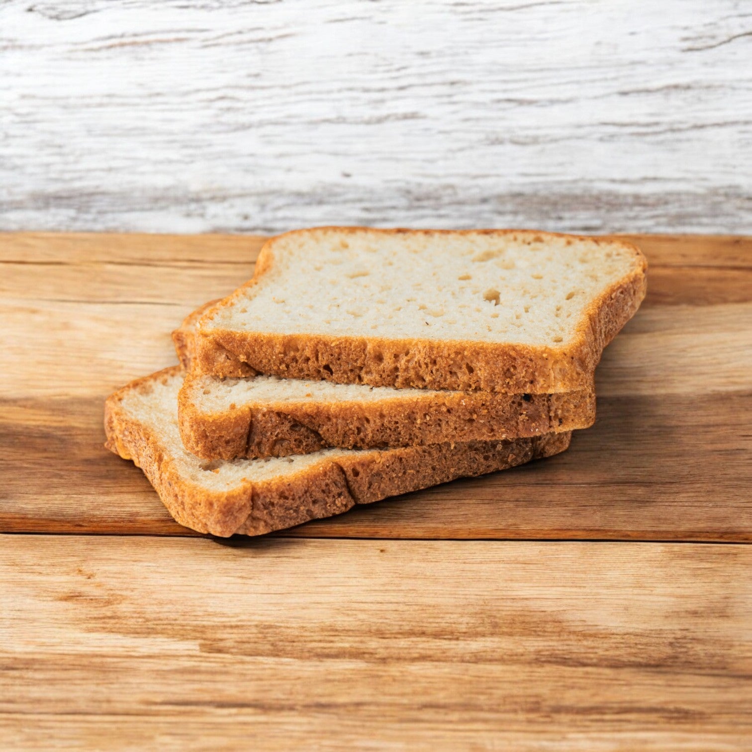 Three slices of OMG It's Gluten Free White Bread Loaf, stacked on a wooden surface against a white wooden background, showcasing an individually sized option with a soft and fluffy texture.