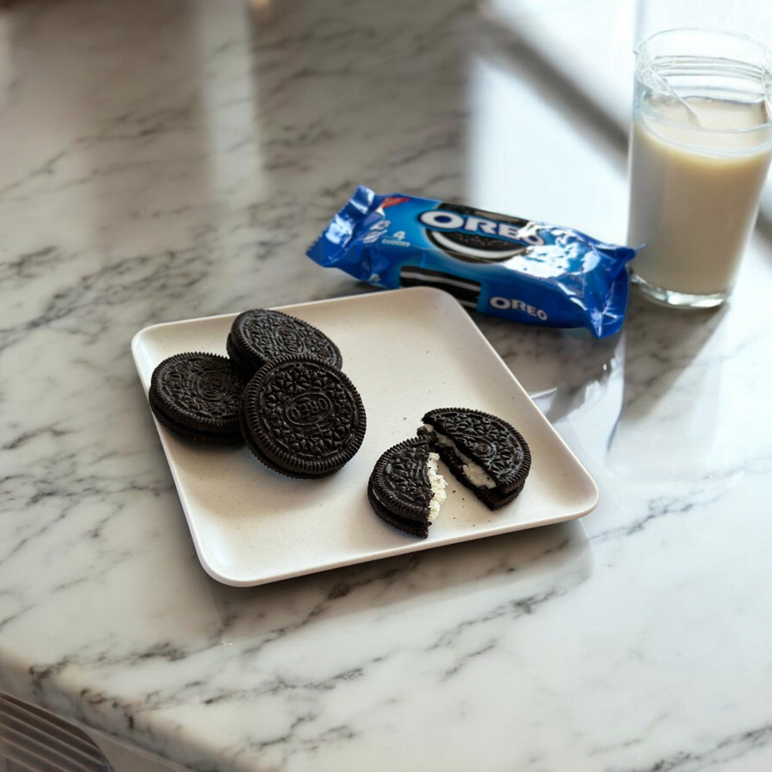 A plate with several OREO Chocolate Sandwich Cookies- 1 Bag, Containing 4 Oreos by Nabisco, and one partially eaten cookie, their chocolate wafers revealing the signature creme filling, sits next to a package of OREOs and a glass of milk on a marble countertop.