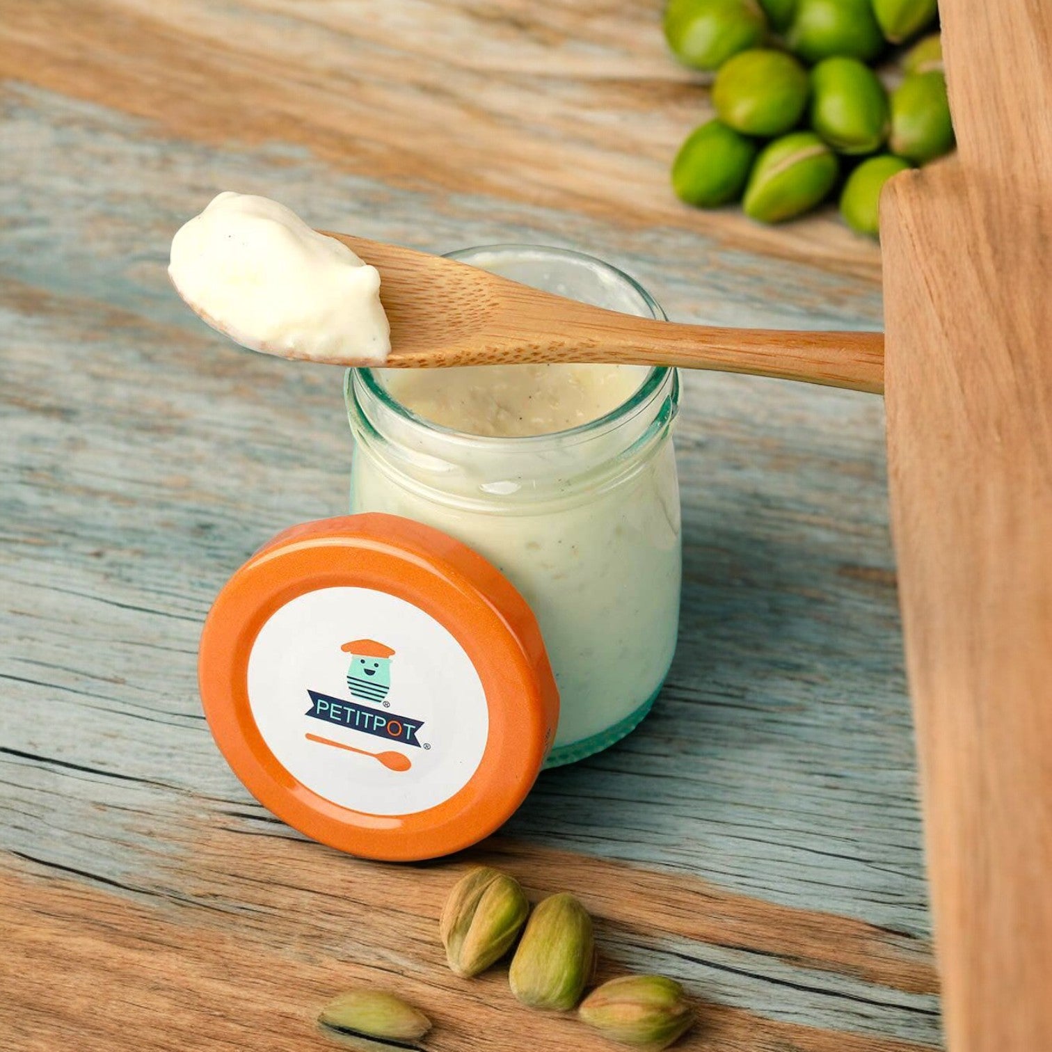 A small glass jar with an orange lid labeled "Petit Pot" containing a creamy white substance. A wooden spoon, filled with the same substance, rests on top of the jar beside some scattered pistachios—exemplifying artisanal French cuisine in this elegant Pistachio Organic French Dessert by Petit Pot.