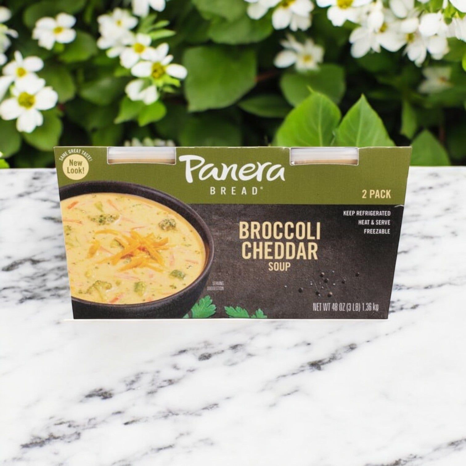 Panera Bread's Broccoli Cheddar Soup 3-pack, each 24 oz, sits on a marble surface adorned with white flowers. This ready-to-heat soup features creamy cheddar cheese for a delightful meal.