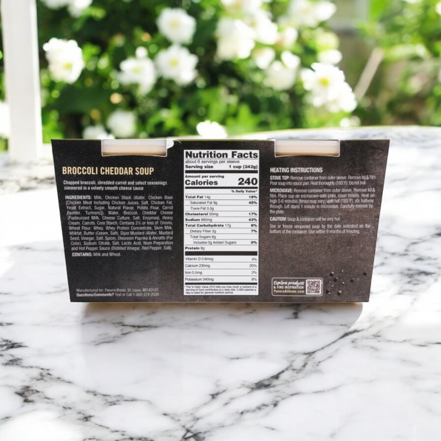 A Panera Broccoli Cheddar Soup – 3 Packs of 24 oz, 2-Count box from Panera Bread sits on a marble surface displaying nutrition facts and ready-to-heat instructions. The aroma of creamy cheddar cheese blends with white flowers in the background.